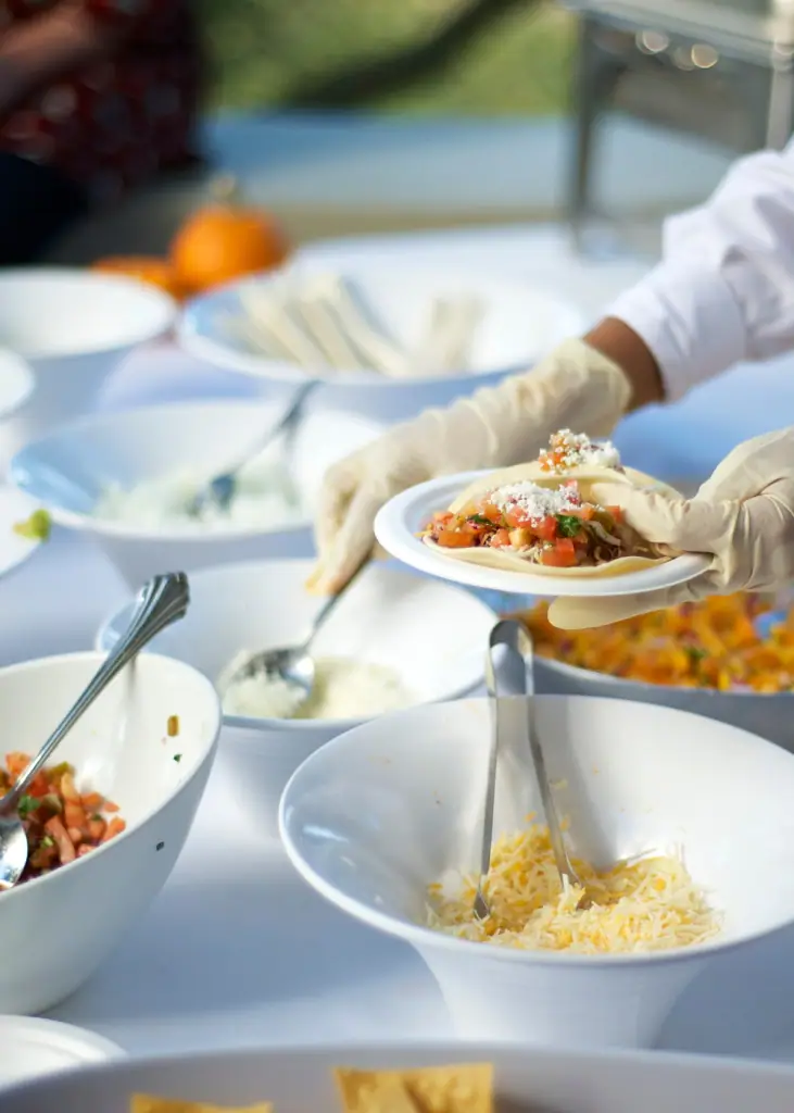 Gloved catering staff plating food at an outdoor buffet with bowls of fresh toppings